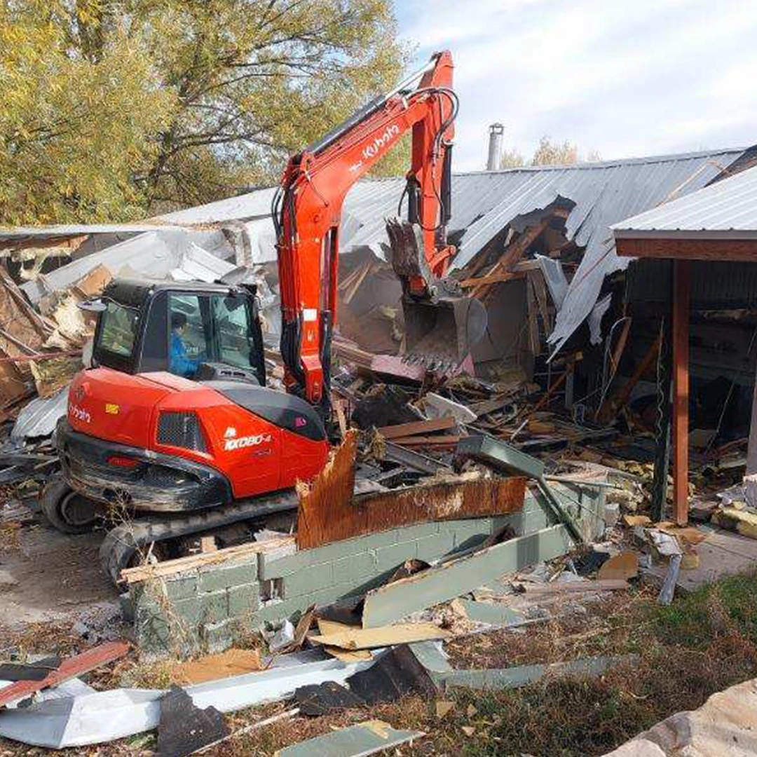 A red excavator demolishes a building, surrounded by debris including wooden beams and metal scraps on the ground. A red excavator demolishes a building, surrounded by debris including wooden beams and metal scraps on the ground.