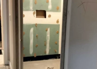 A view through a doorway into a room under construction, with light green drywall sections installed on the wall, marked with circular and vertical adhesive applications.