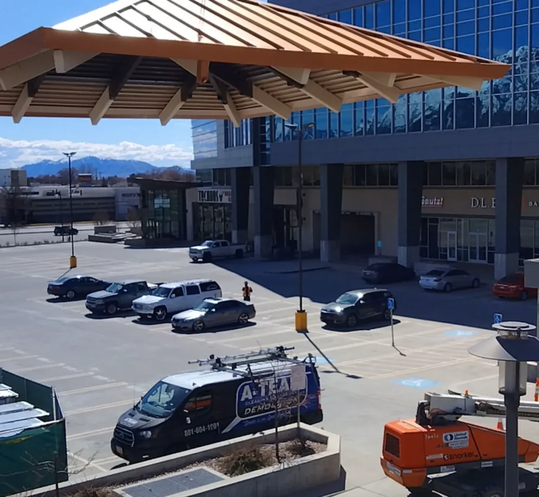 A wide shot of a parking lot in front of a modern glass building with a distinct wooden awning, showing several parked cars, a black van with a "A-TEAM DEMO" logo, and a small orange lift vehicle. Mountains are visible in the background. A wide shot of a parking lot in front of a modern glass building with a distinct wooden awning, showing several parked cars, a black van with a "A-TEAM DEMO" logo, and a small orange lift vehicle. Mountains are visible in the background.